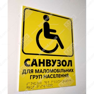 Тактильна табличка жовтого кольору з текстом санвузол для маломобільних груп населення та шрифтом Брайля фотографія ліворуч